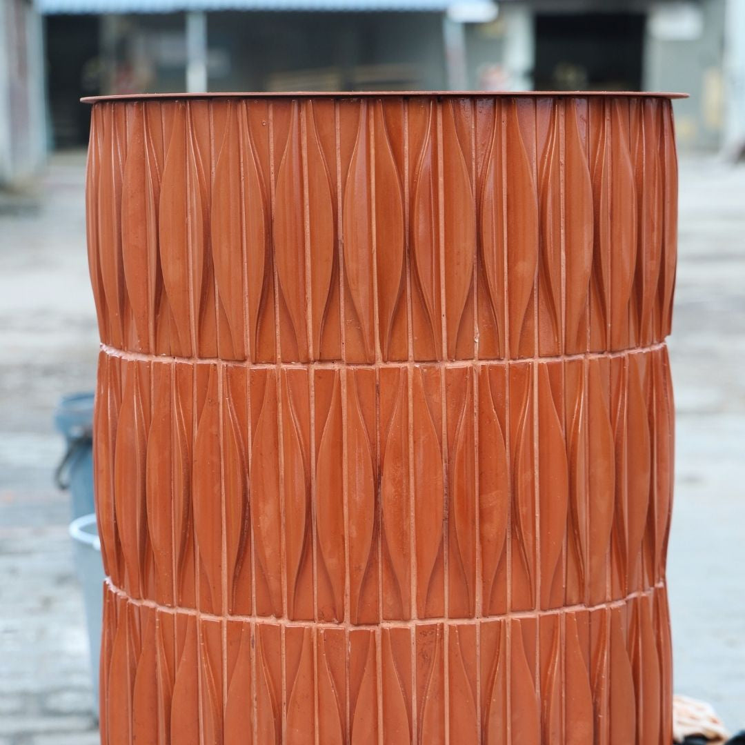 
                  
                    A cylindrical barrel-shaped pillar with a textured, vertically ribbed surface and a flat top in a terracotta orange color, sitting in an outdoor workshop setting.
                  
                