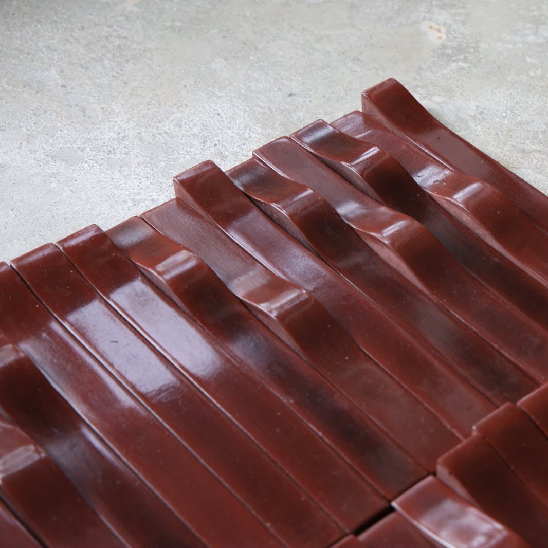 
                  
                    A close-up of several dark reddish-brown textured tiles arranged diagonally on a light gray concrete-like surface.
                  
                