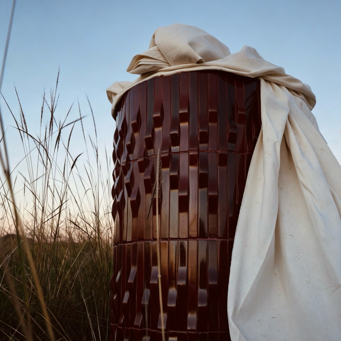 
                  
                    A cylindrical outdoor sculpture or planter made of dark reddish-brown textured panels, draped with a cream fabric, photographed outdoors in a grassy field at sunset.
                  
                