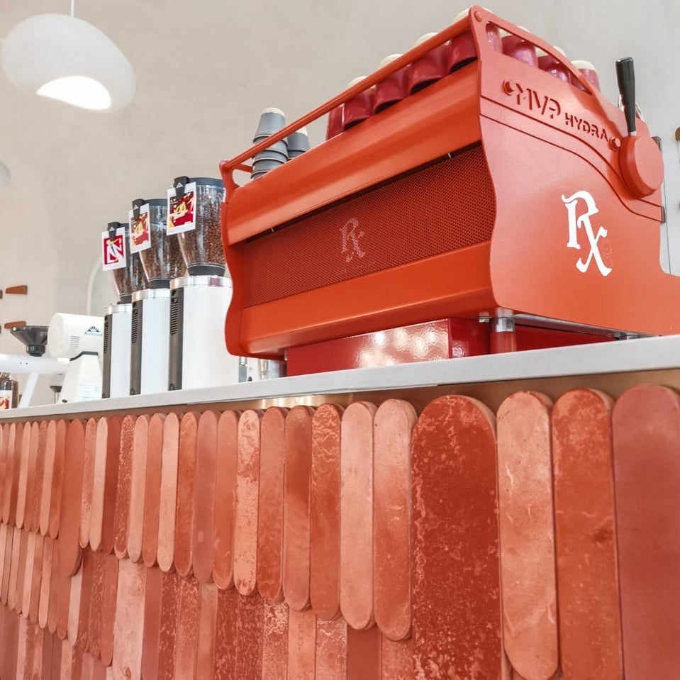 
                  
                    Red espresso machine with a white logo on a bar counter with decorative wall panels.
                  
                