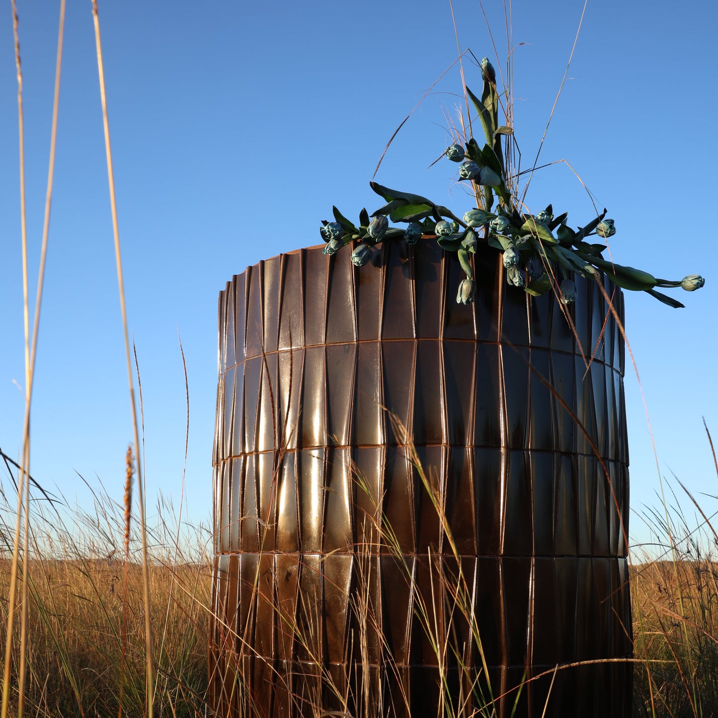 
                  
                    A tall, cylindrical pillar of vertically stacked bronze-toned tiles, placed in a field of dry grass under a clear blue sky, with a small green plant placed on top.
                  
                